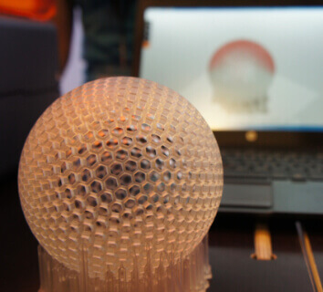 SLS printed 3D sphere with hexagonal structure and supports, 3D image file on laptop screen in background